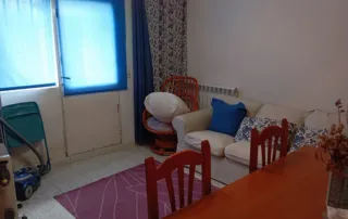 a sala de estar y el comedor de una vivienda. Se puede observar un sofá, una alfombra, una mesa de comedor con sillas y una aspiradora