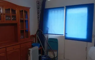 habitación con un mueble de madera, dos persianas azules en las ventanas, una aspiradora, un radiador eléctrico y un cubo azul en el suelo