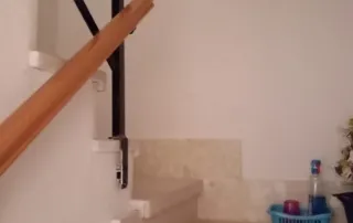 la parte inferior de una escalera en una vivienda, con un pasamanos de madera y barandillas de metal