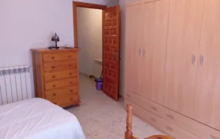 interior de un dormitorio, cómoda de madera con cinco cajones, armario grande de seis puertas, cama con sábanas blancas y una cabecera de madera