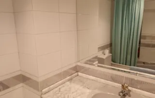 cuarto de baño con azulejos blancos, un espejo de pared a pared, un lavabo con encimera de mármol y una cortina de ducha de color verde azulado.