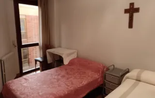 muestra el interior de una habitación con dos camas individuales, una mesa pequeña y una cruz en la pared.