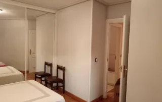 el interior de una habitación con dos camas individuales, dos sillas de madera, y un armario con puertas de espejo