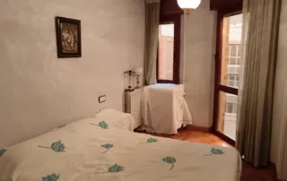 habitación de un apartamento o casa, contiene una cama, un radiador, una ventana y un cuadro en la pared.